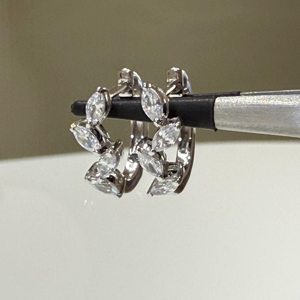 May include: A pair of white gold hoop earrings with a leaf design. The earrings are made of metal and have small, clear stones that resemble diamonds. The earrings are shown on a white background.